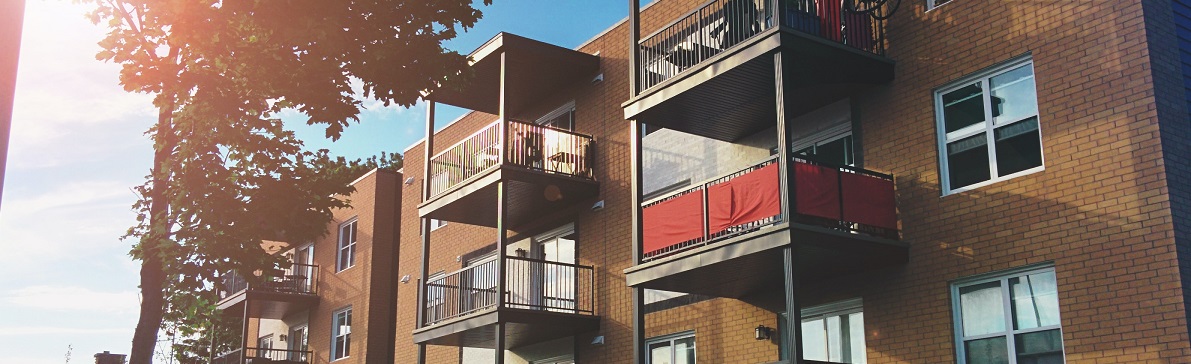 The outside of an apartment building with a couple balconies featuring tall trees and the sun shining intensely. 