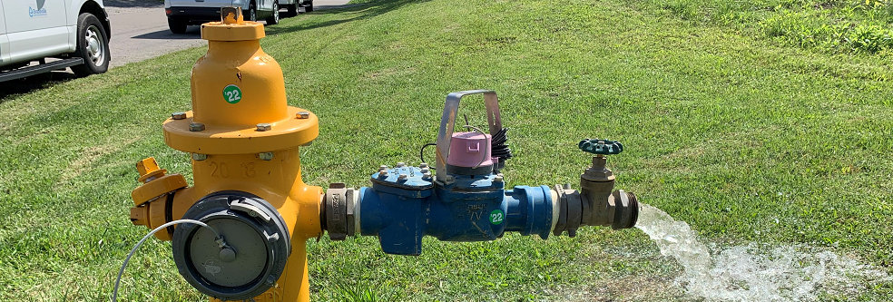 A yellow fire hydrant in a lawn with an attached hydrant meter with water flowing out. 