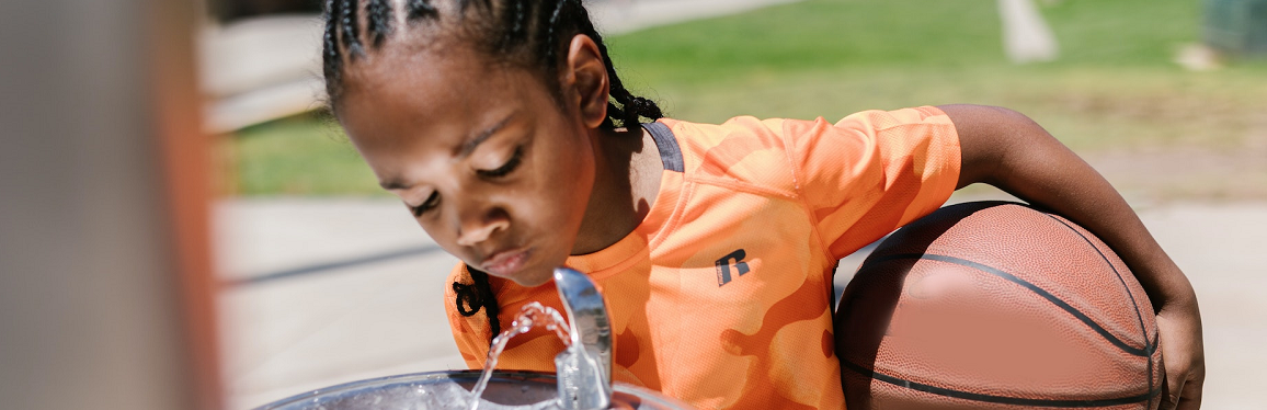 A young boy in an orange shirt holding a basketball while drinking from an outdoor water fountain. 