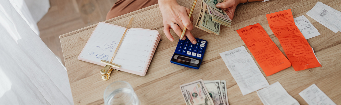 An overhead view of a person hold a stack of cash and a pencil while typing on a calculator. A calendar planner, money, and receipts are scattered on the table implying the person is calculating their bills.