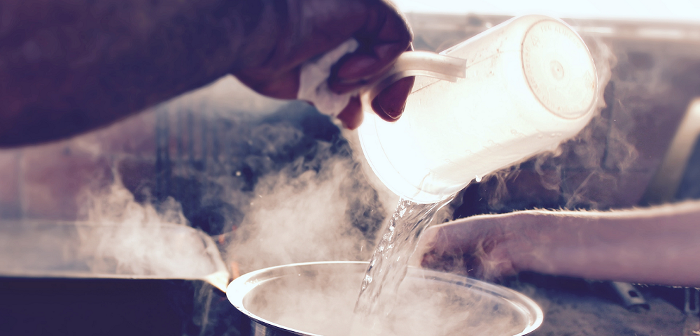 A person pours boiling, hot, steaming water into a steel pot. 