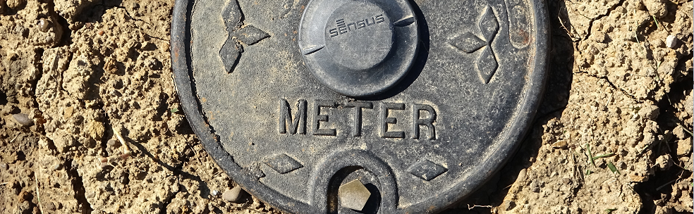 A close up of a black water meter in a dirt ground.